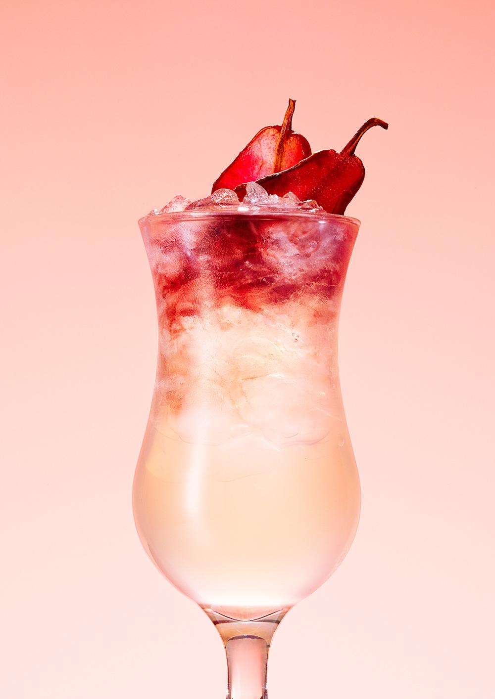 A cocktail glass containing a pale yellow drink with a red layer on top, garnished with two red pomegranate or apple-like fruit slices.