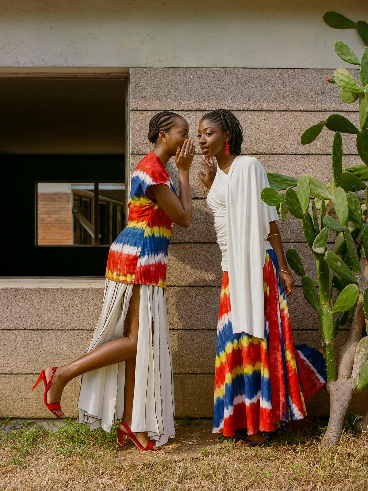 Two women in tie-dye clothing stand facing each other in front of a wall.