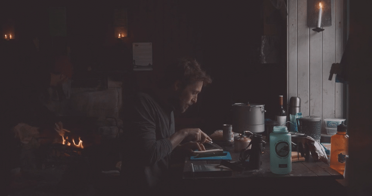 A man is cooking in a dimly lit, rustic kitchen with a fire burning in the background.