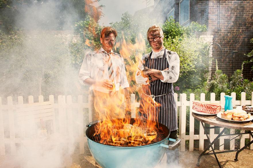 Two men, one in a chef's white shirt and the other in a black and white striped apron, standing in front of a large, blue, flaming barbecue grill.
