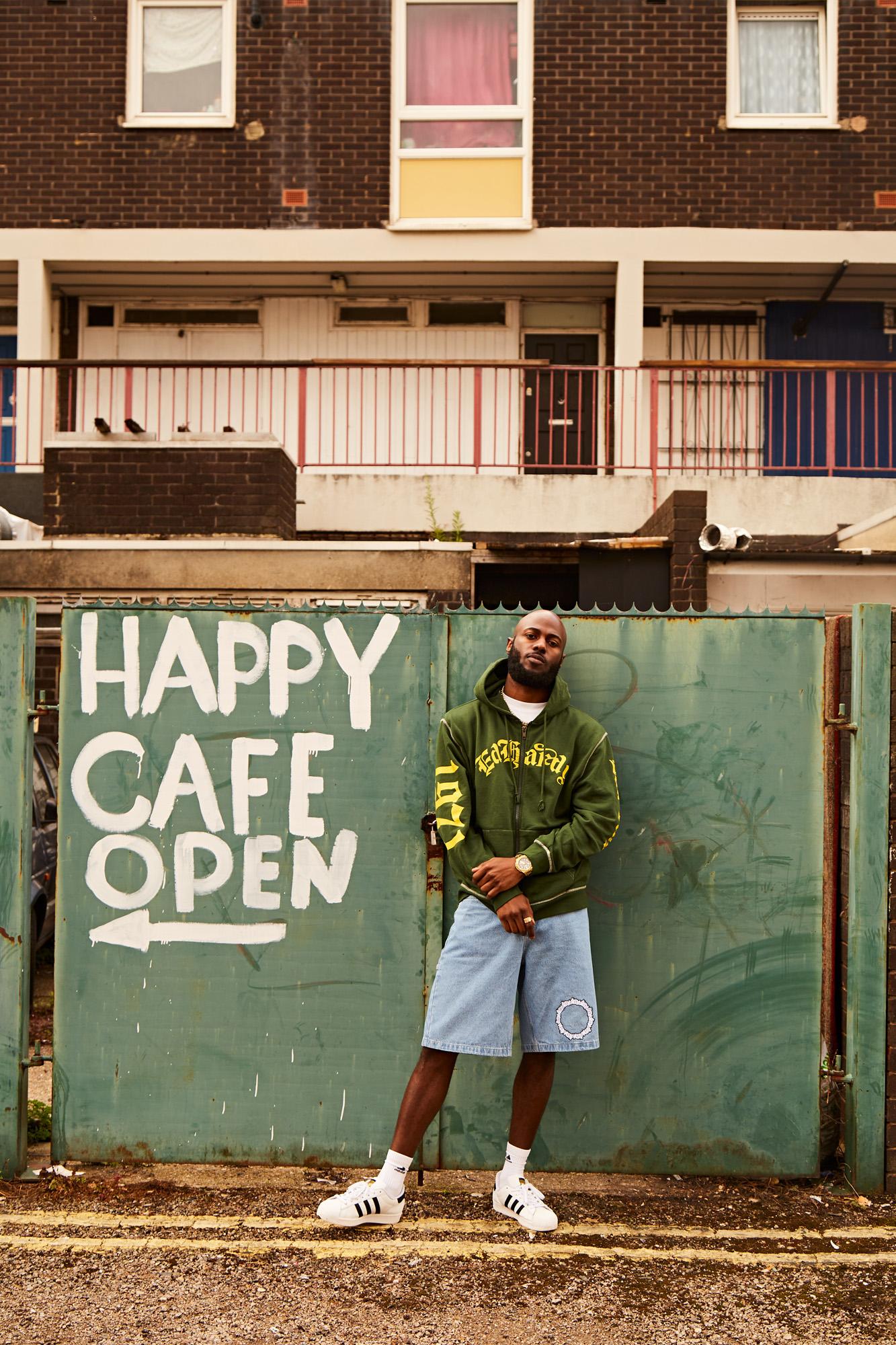 A man in a green hoodie and denim shorts stands in front of a graffiti-covered wall with a sign that reads 'Happy Cafe' in a rundown urban area.