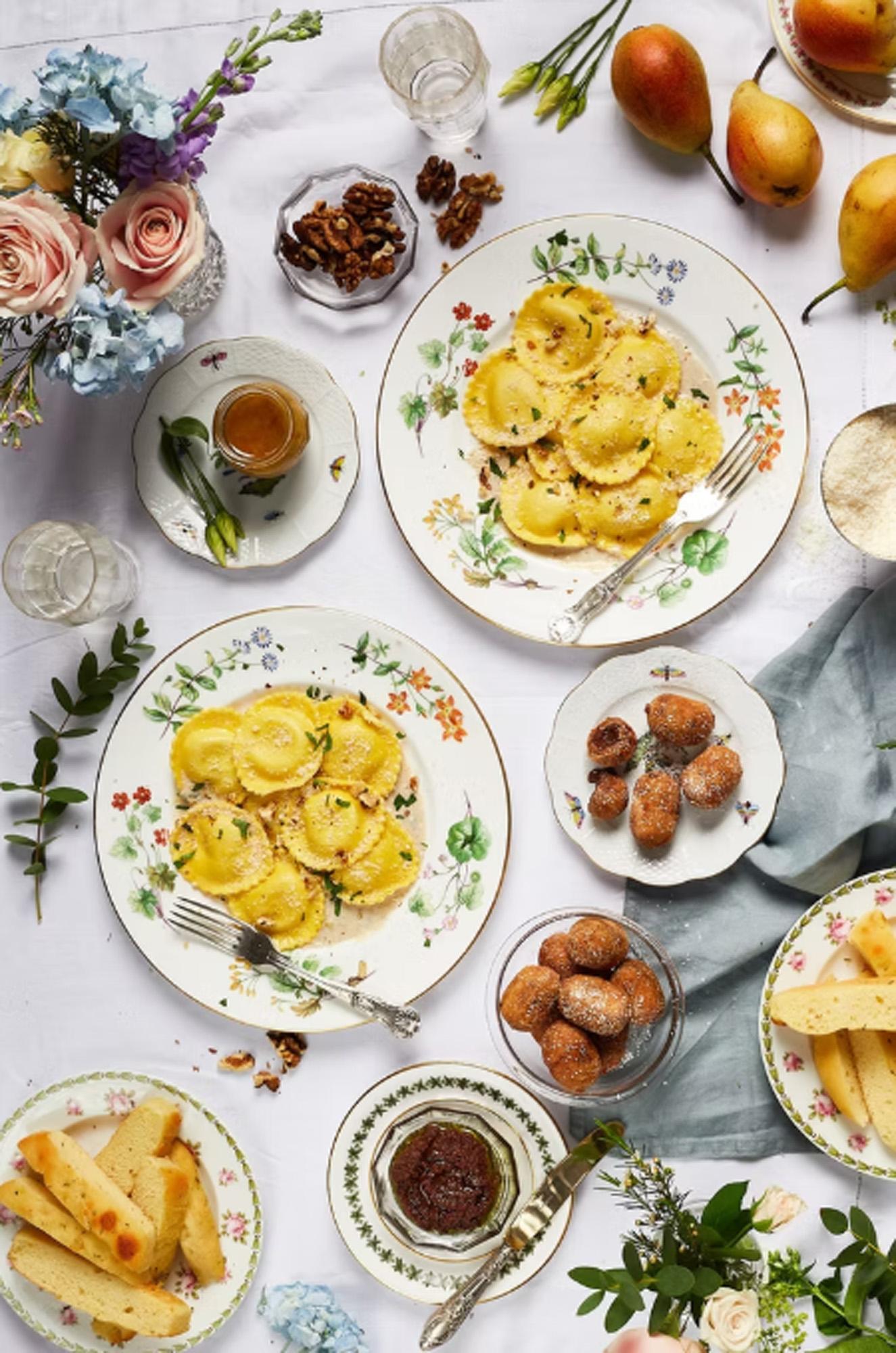 A table setting with Italian dishes like ravioli and pasta on a white tablecloth with flowers.