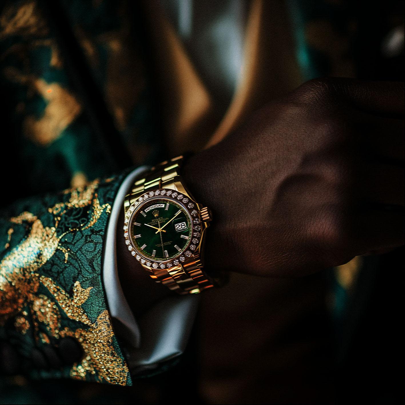A gold Rolex watch with a green face and diamond-encrusted bezel is being worn on a dark-skinned wrist.