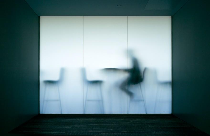 A dimly lit, empty room with a large, white wall featuring a haunting, blurred image of a person sitting in a chair, with a faint, ghostly outline of a cityscape in the background.