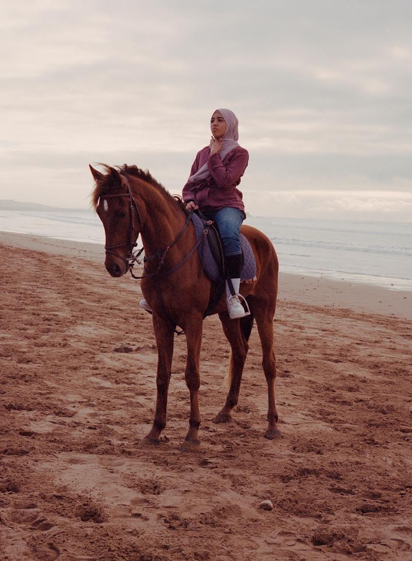 A woman in a hijab riding a horse on a beach, wearing a red jacket, blue jeans, and white sneakers.