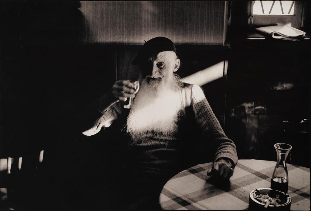 An elderly man sits in a chair holding a glass of wine.