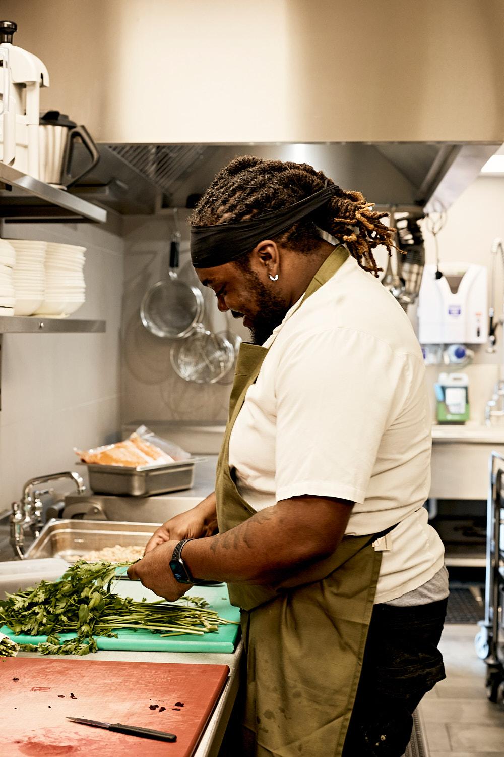 A chef in a white chef's coat and black hat is chopping fresh herbs in a commercial kitchen.