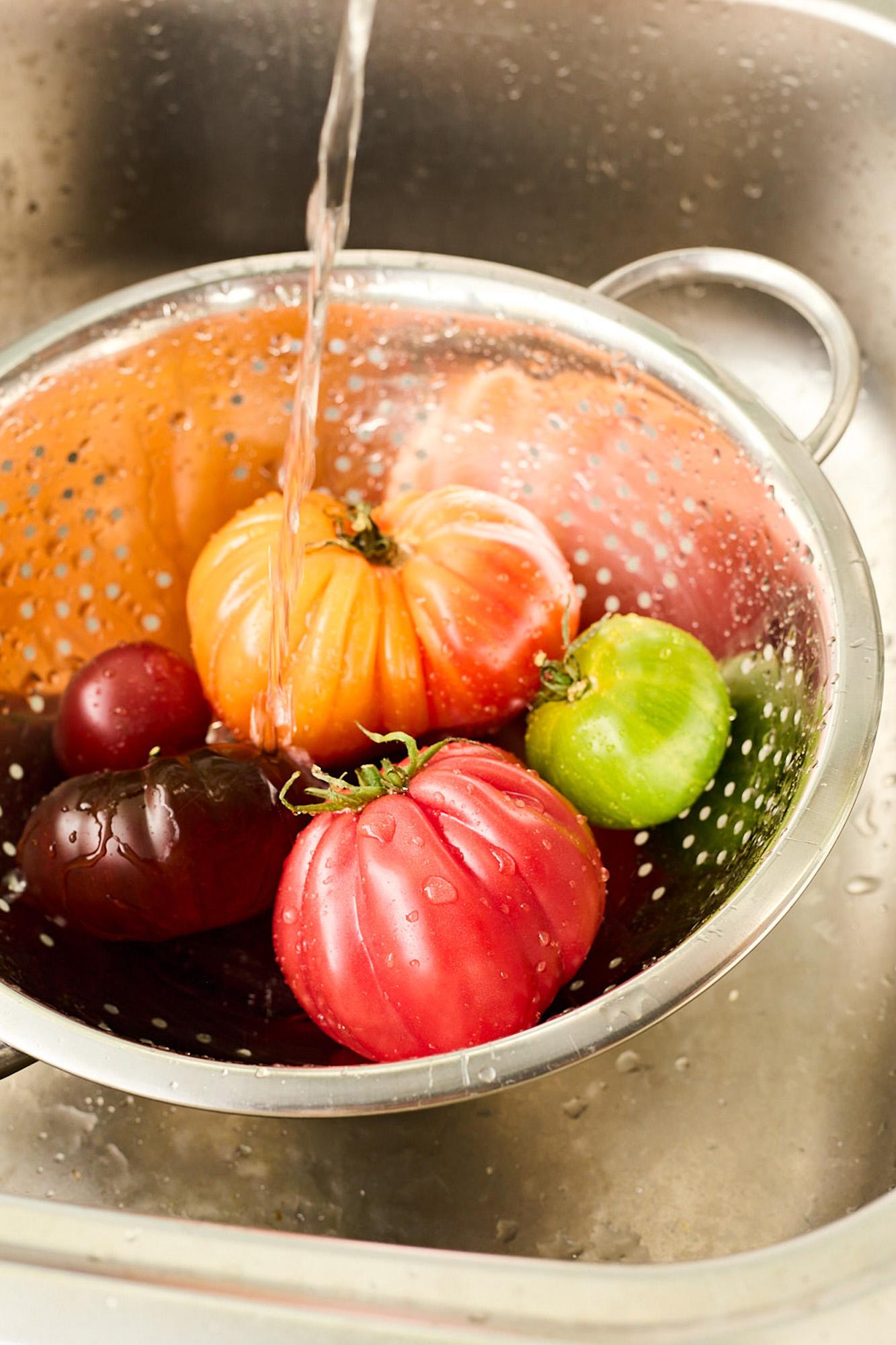 A stainless steel colander filled with fresh, colorful tomatoes, including red, green, and yellow varieties, being washed under a stream of water.