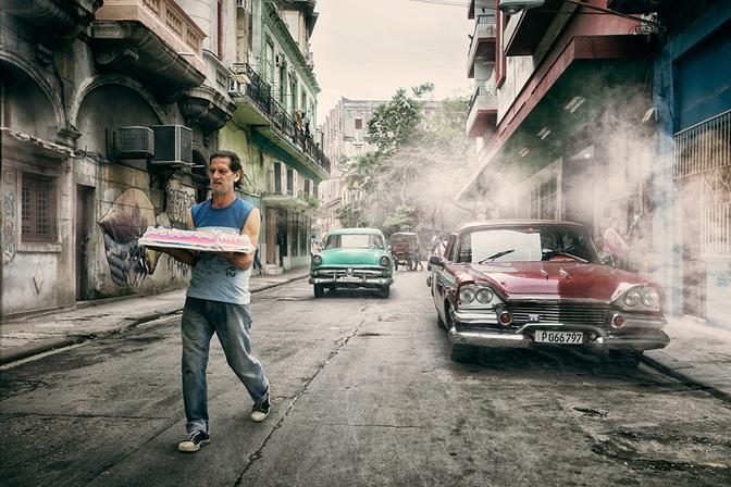 A man in a blue shirt and jeans is walking down a street in Havana, Cuba, carrying a large tray of pastries.