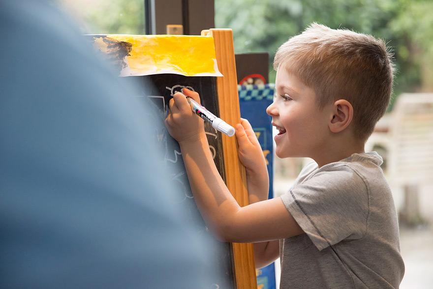 A young boy with short, blond hair is drawing on a blackboard with a white marker.