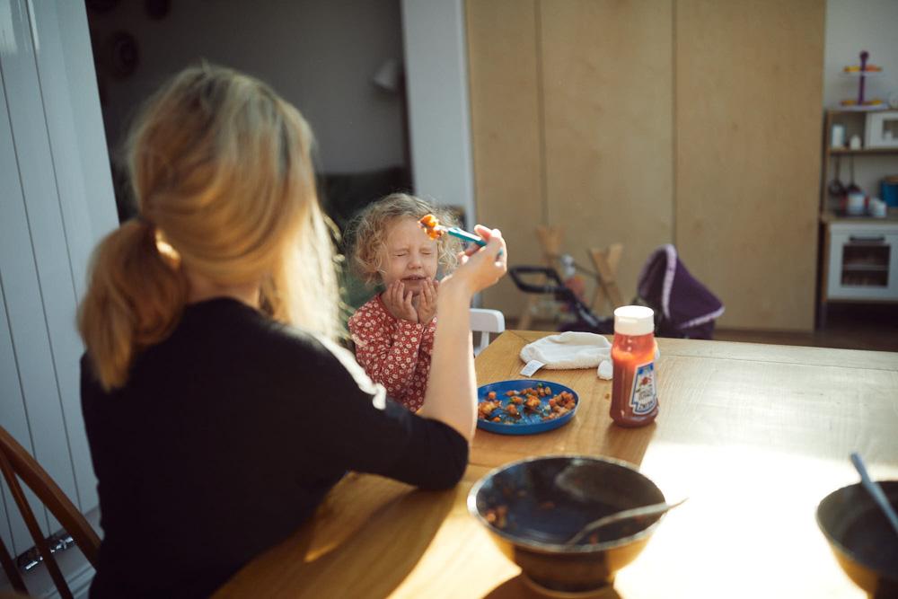 A woman with blonde hair in a ponytail sits at a table with a young girl. The girl is eating a spoonful of food, while the woman watches with a gentle, concerned expression.