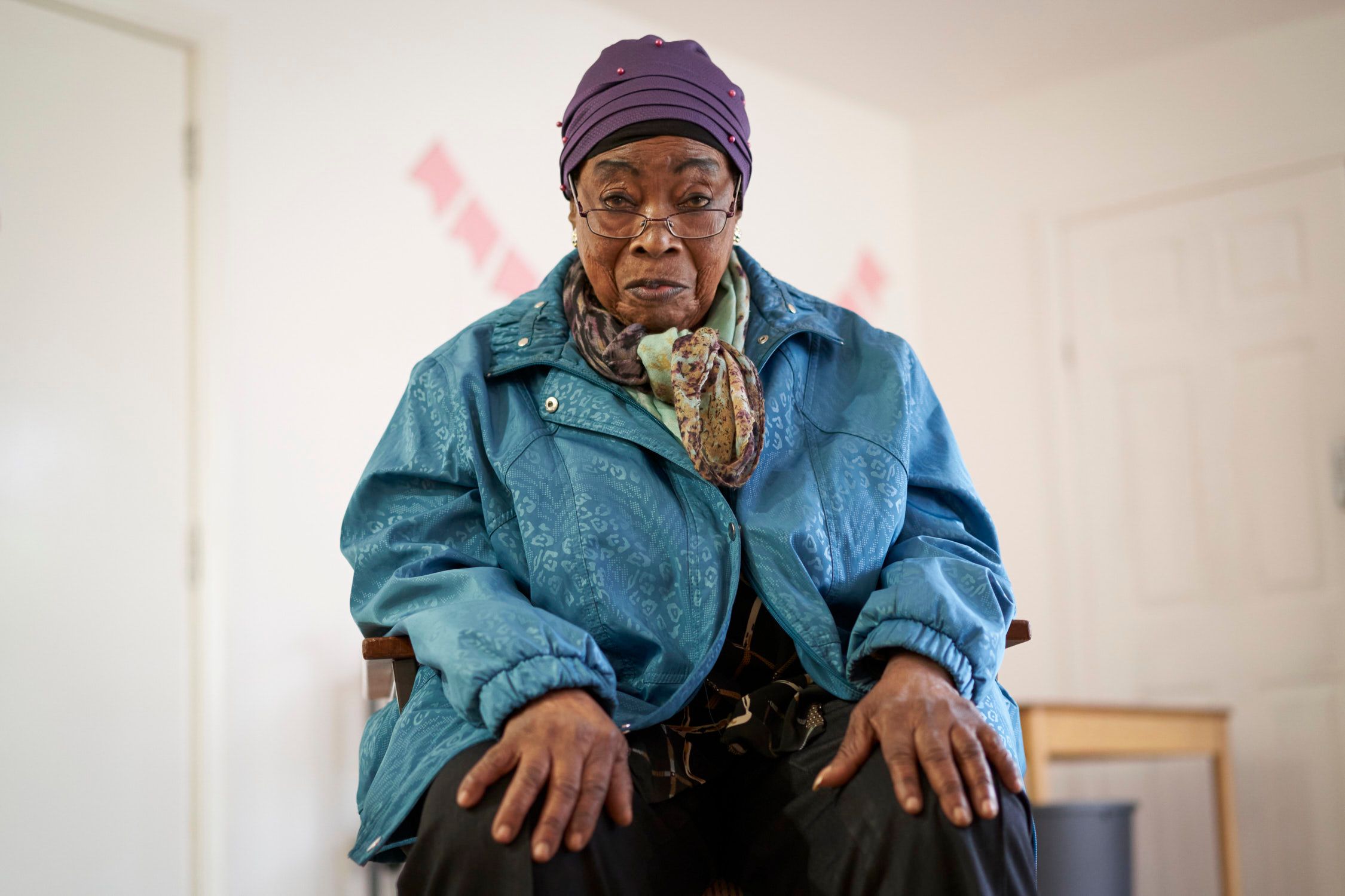 An elderly woman sitting in a chair, wearing a blue jacket, a purple hat, and glasses, in a white room with a door and a window in the background.