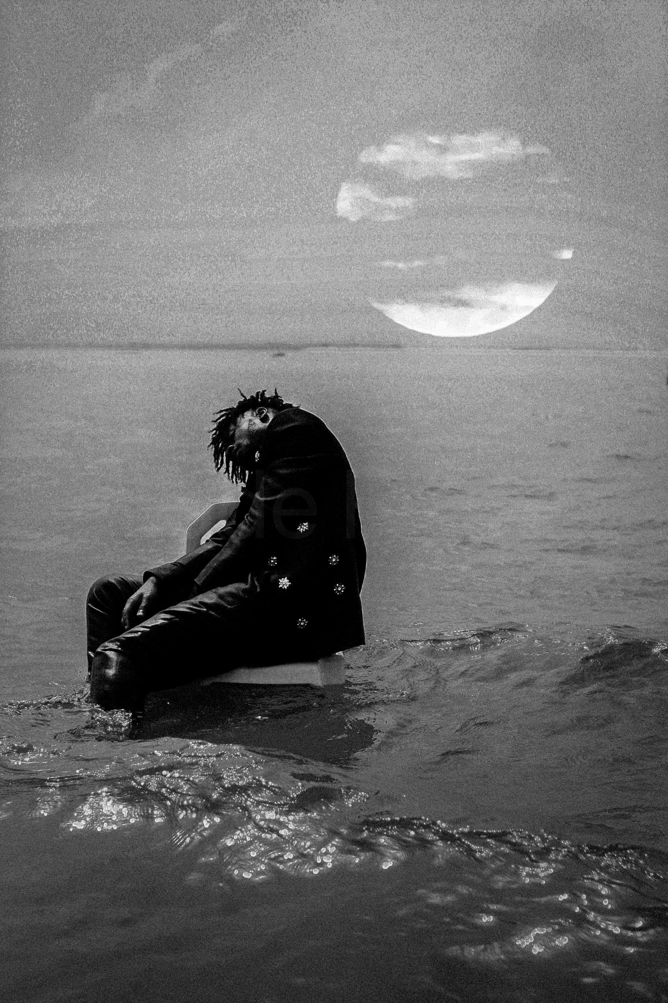 A person sitting on a chair in the water, wearing a black suit and no shoes.