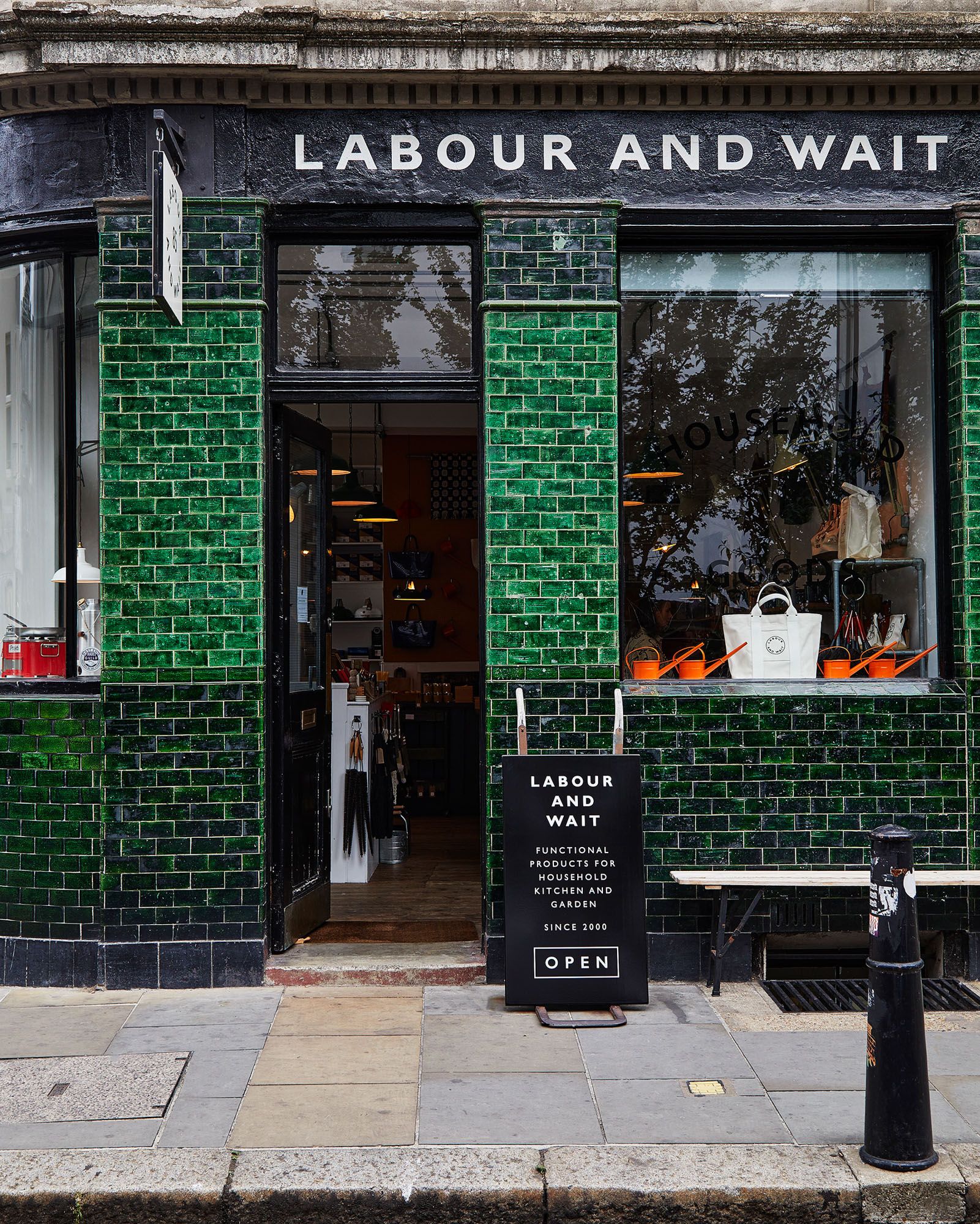 A storefront with a green brick facade and a sign reading 'Labour and Wait' above the entrance, with a window display of various household and kitchen items.