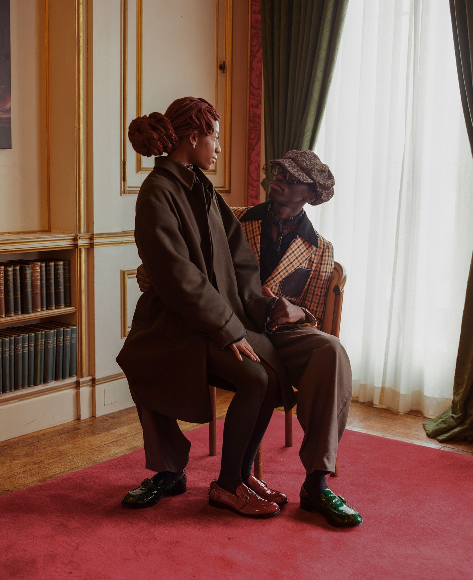A man and a woman sit on a chair in a room with a bookshelf and a window, dressed formally.