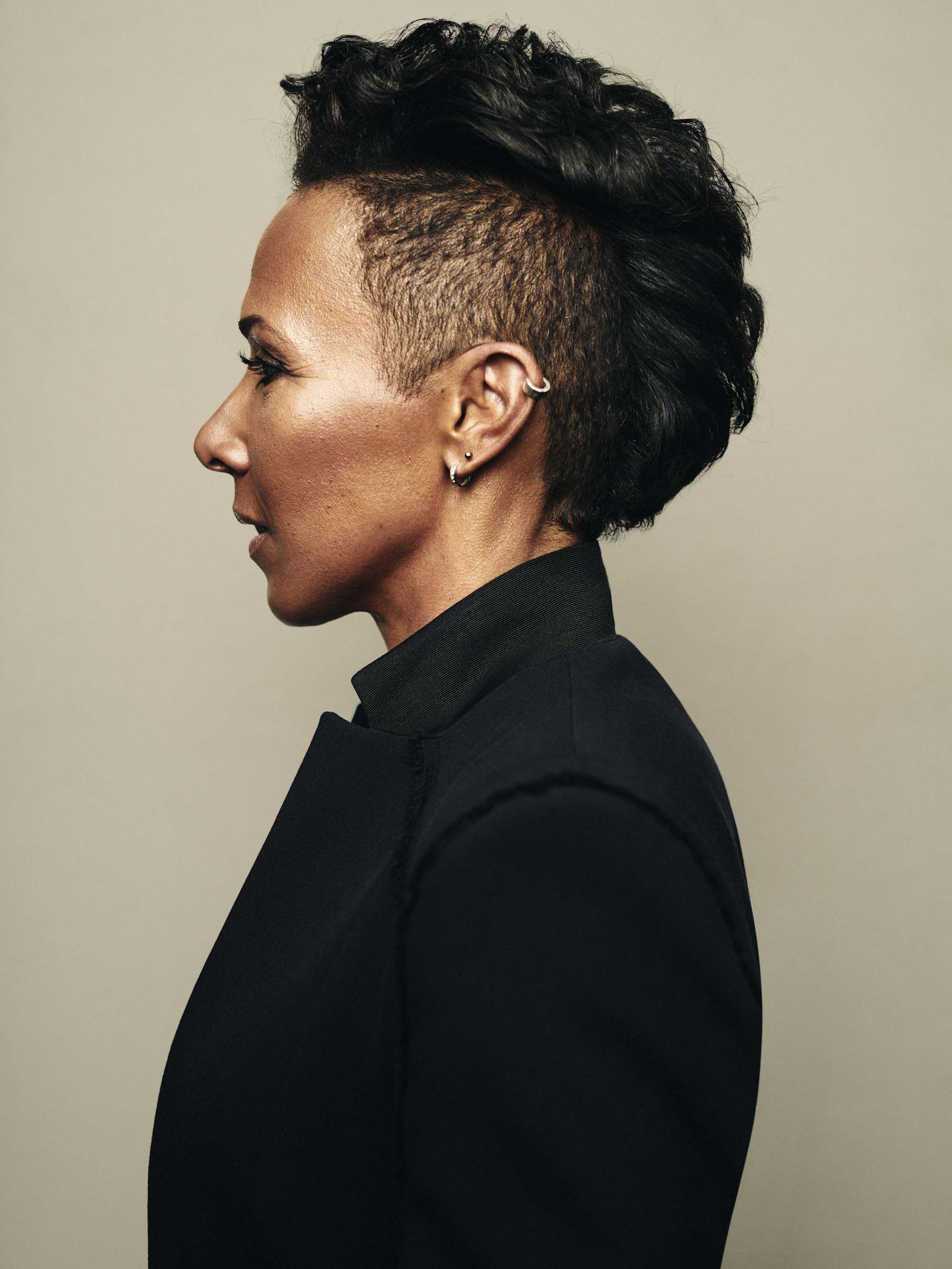 A woman with a shaved head and a short, spiky hairstyle, wearing a black leather jacket and a silver earring.