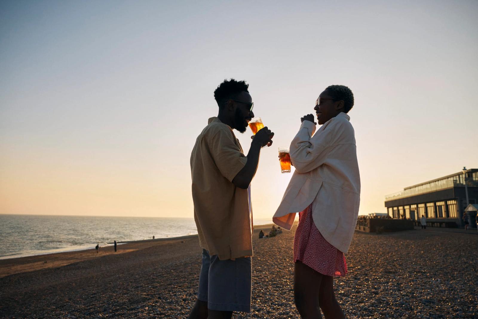 A man and woman stand on a beach, each holding a glass of beer, facing each other in a relaxed, casual manner. The man is wearing a tan shirt and shorts, while the woman is.