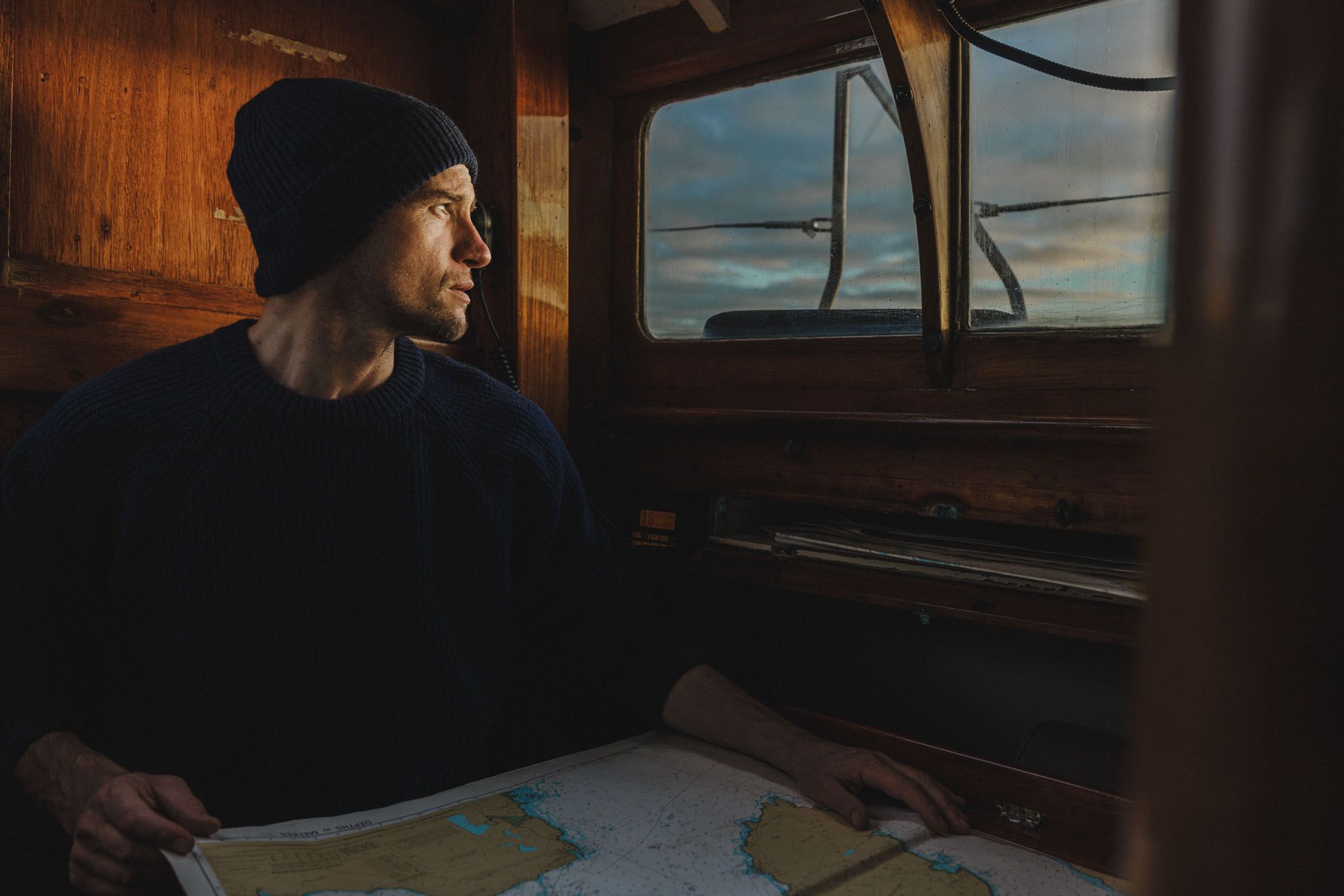 A man in a navy blue sweater and beanie, holding a nautical chart, sitting in a wooden cabin with a large window overlooking the ocean.