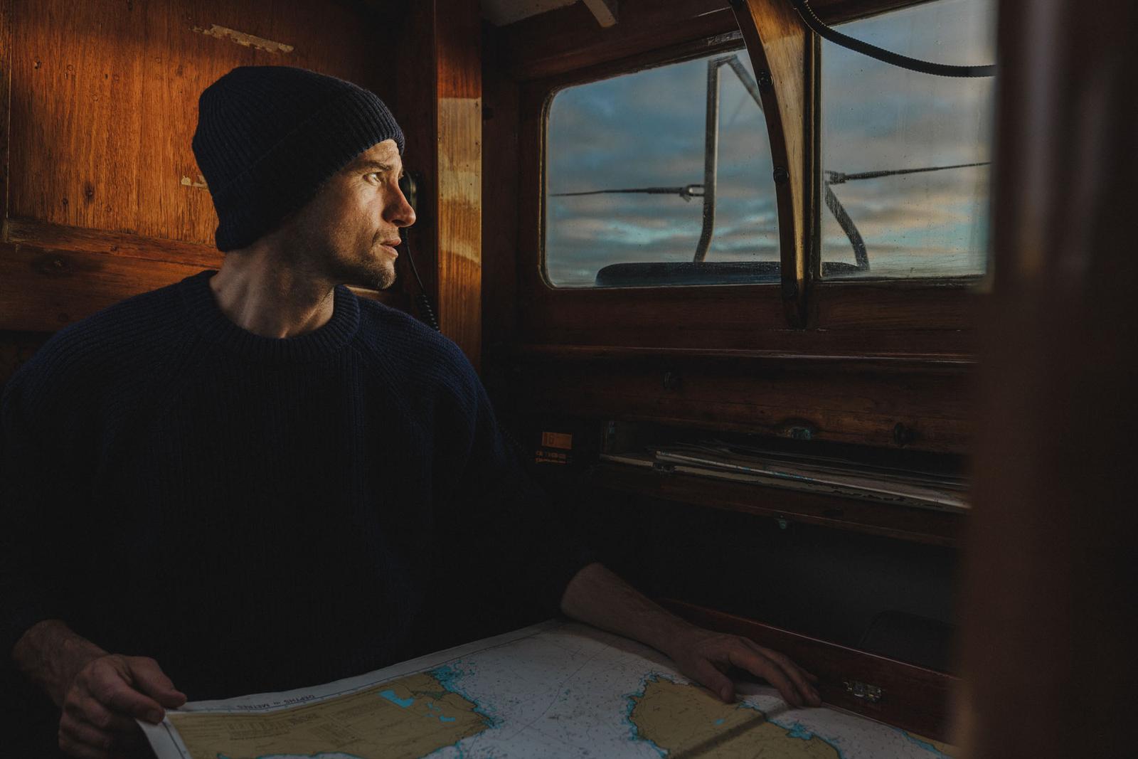 A man in a navy blue sweater and beanie, holding a nautical chart, sitting in a wooden cabin with a large window overlooking the ocean.