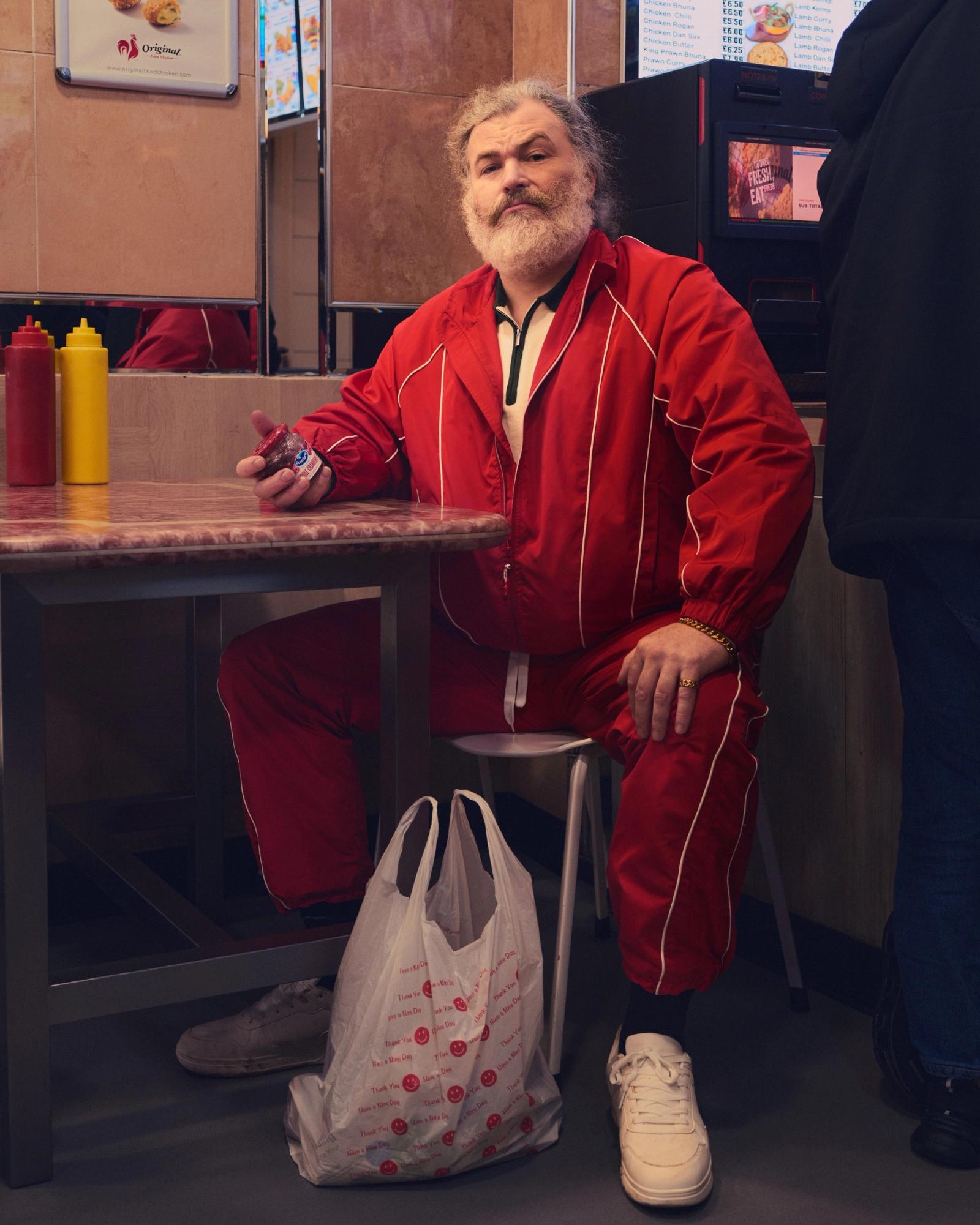 A man in a red and white striped suit, with a white beard and hair, sitting at a table in a fast food restaurant, holding a bottle of special sauce.