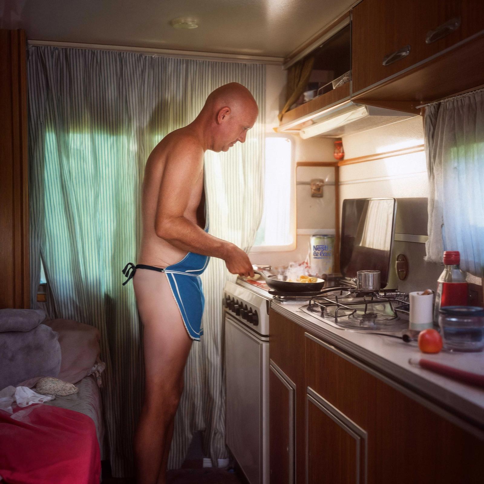 The image shows a side profile of a naked, bald man cooking at a small stove inside a vintage-style camper van. He is wearing only a blue-and-white patterned apron that is tied around his waist. Steam rises from a frying pan as he works in the compact kitchen area, which is filled with soft, natural light from a nearby window.