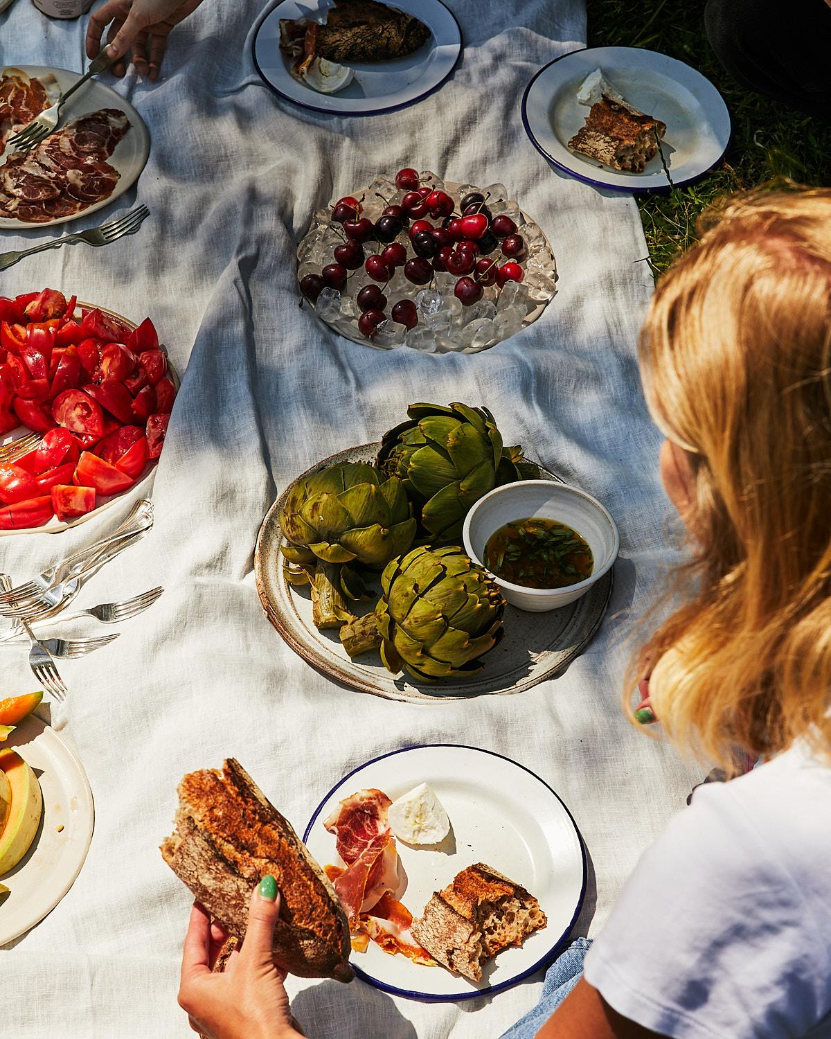 A woman is holding a slice of bread in a picnic setting with a variety of food and drinks, including artichokes, bread, cheese, fruit, and wine.