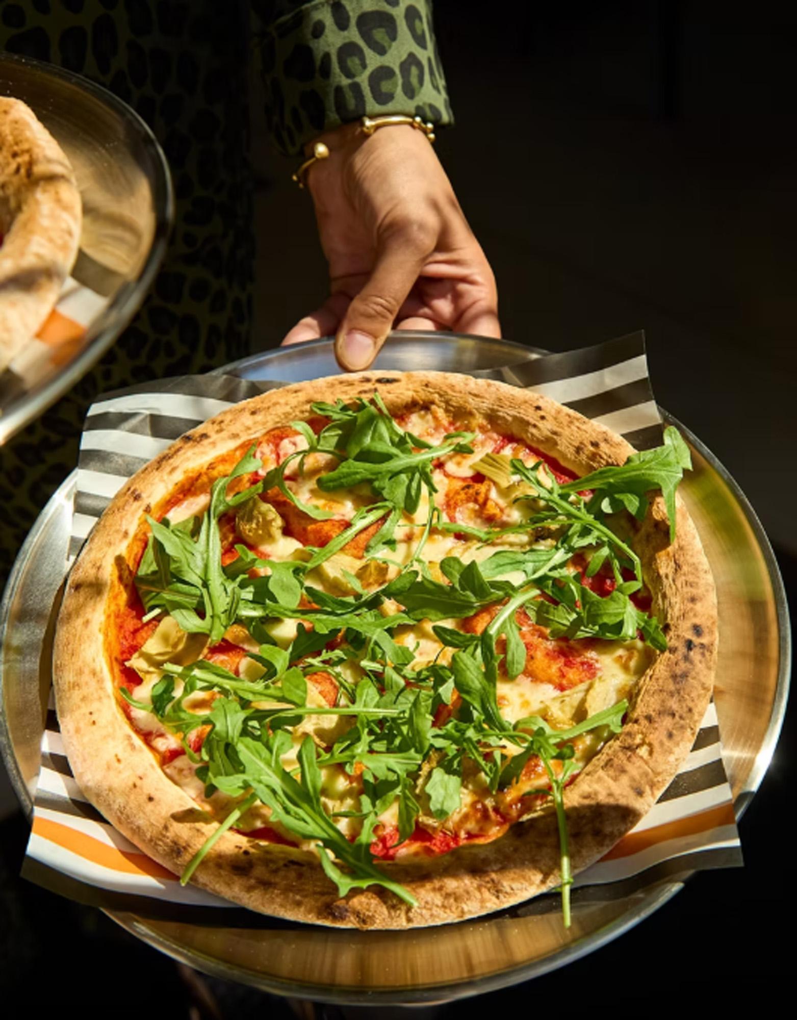 A person holding a pizza with various toppings, including arugula, on a plate.