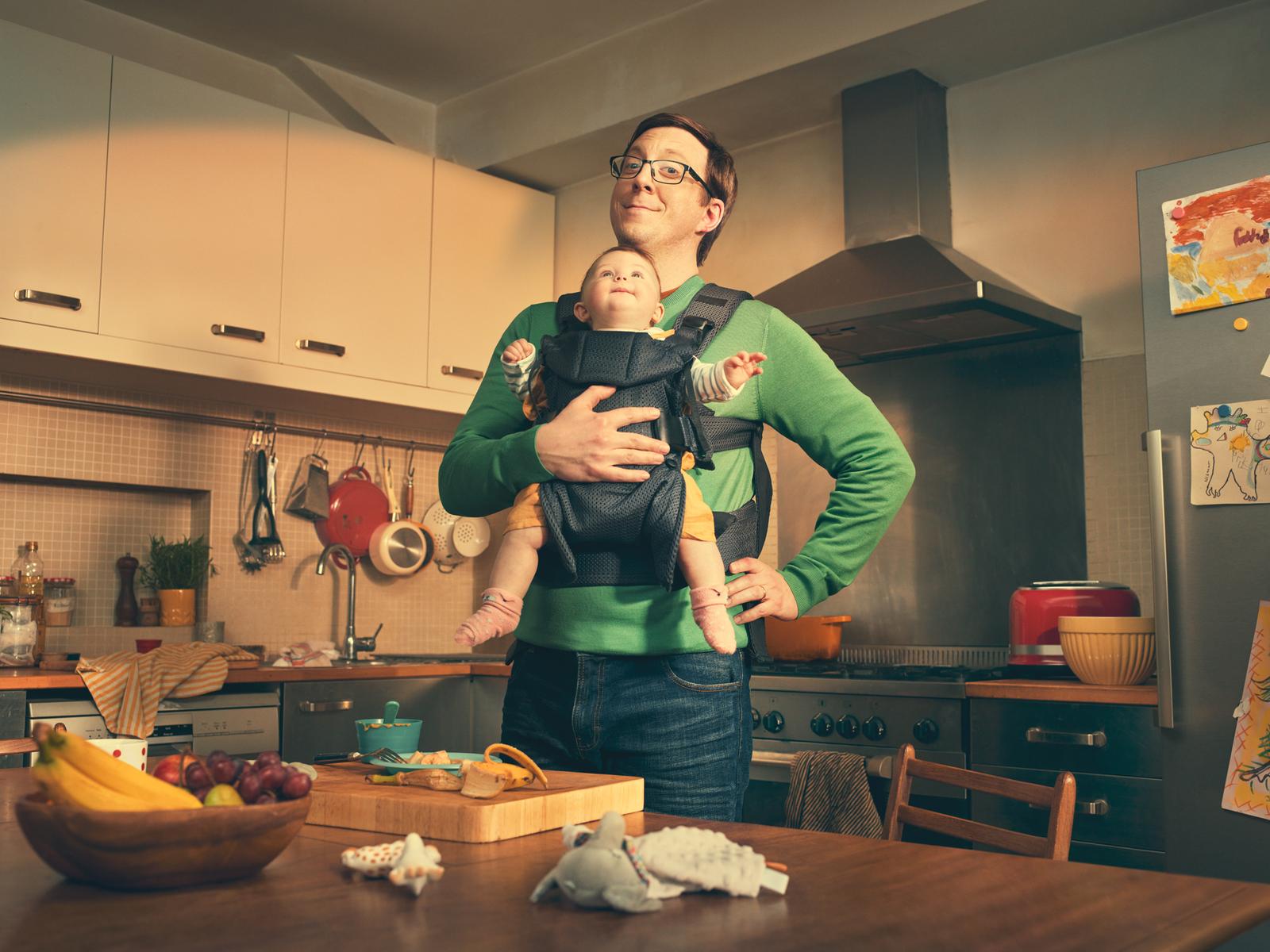 A man in a green sweater and jeans holding a baby in a black baby carrier, standing in a kitchen with a wooden table and various kitchen utensils and food items.
