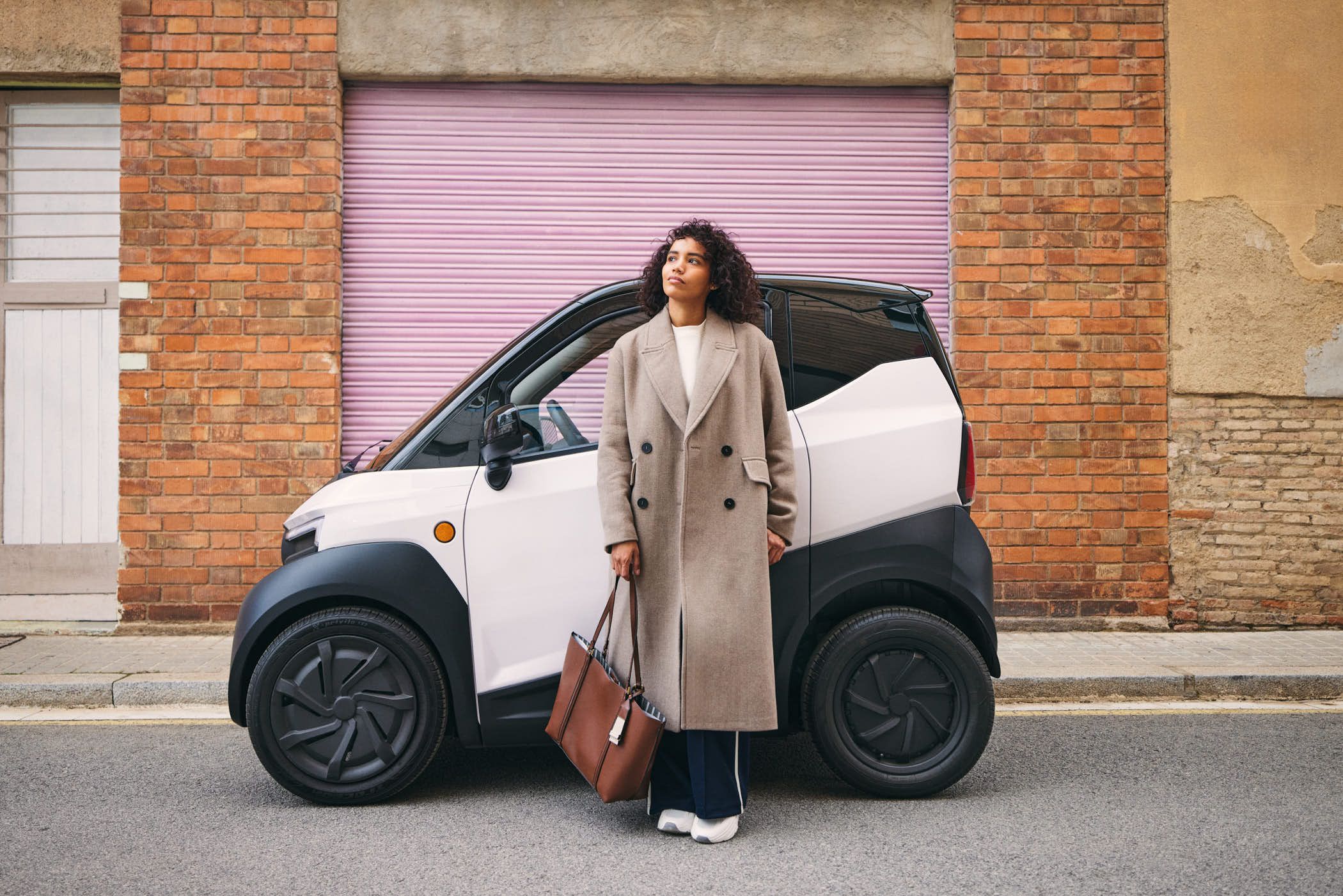 A woman in a long, beige coat stands in front of a white electric car, looking up at the sky with a thoughtful expression. The car is parked on a street in front of a brick.