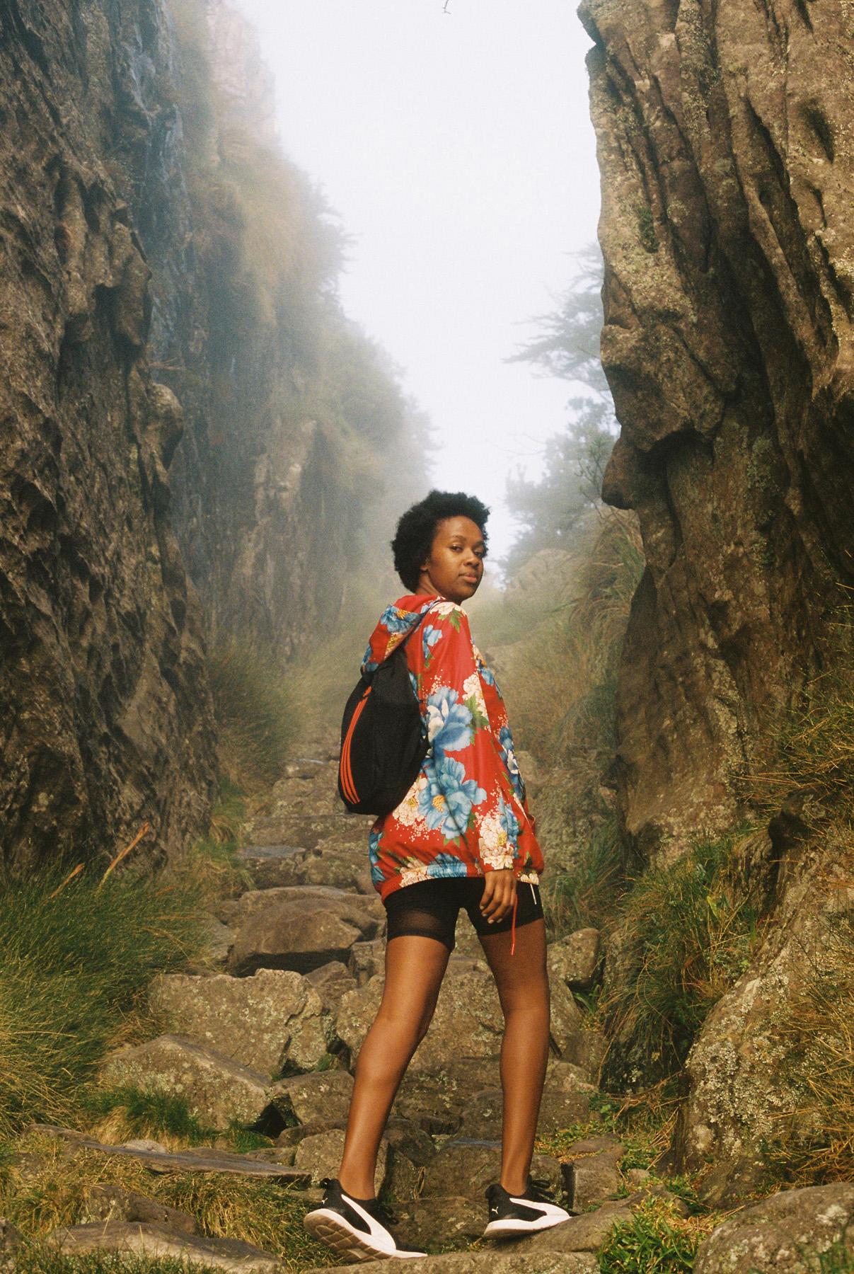 A woman with short, dark hair and a red jacket with blue and white flowers, standing on a rocky outcrop, looking out at a misty landscape.