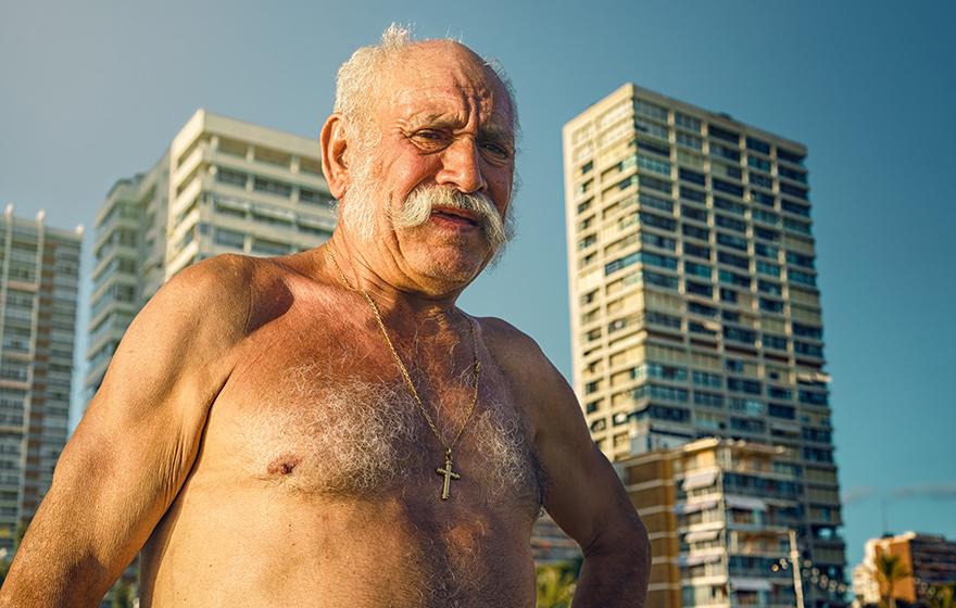 A man with a white beard and mustache, wearing a pair of sunglasses and a beach towel, standing in front of a city skyline with high-rise buildings in the background.