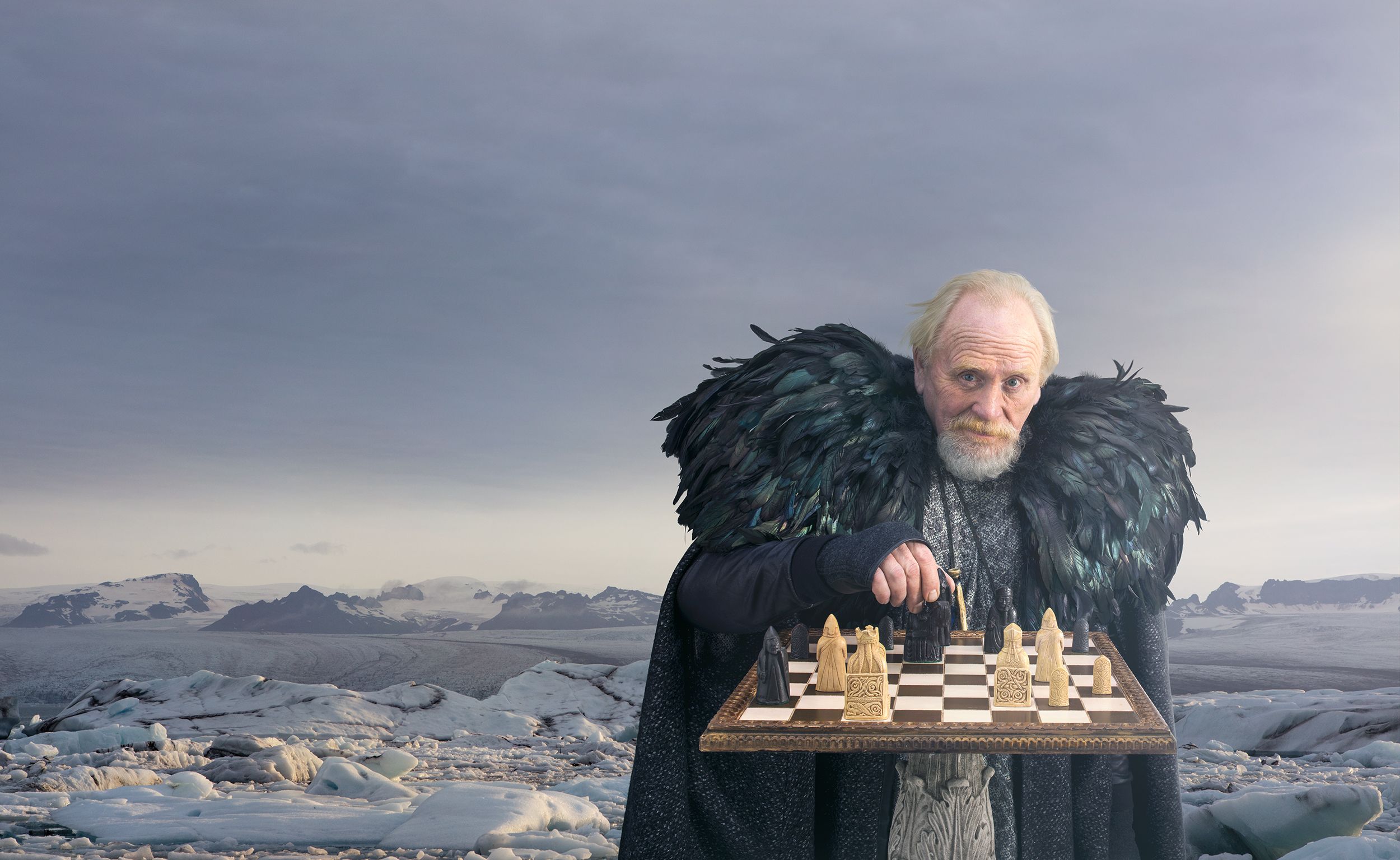 A man in a black feathered cape and a grey tunic stands on a glacier, holding a chessboard with a piece in his hand.
