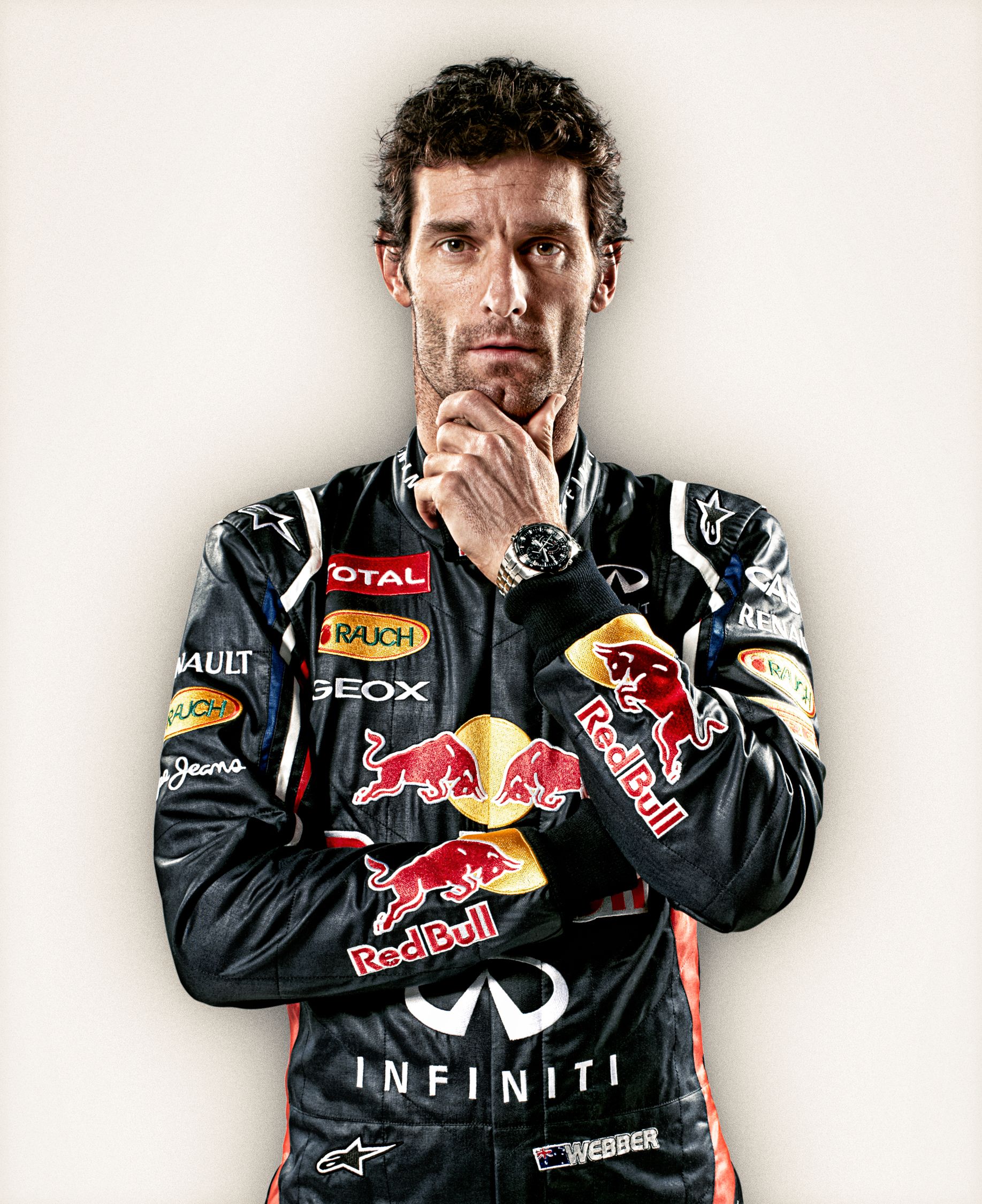 A man in a black racing suit with logos of Red Bull, Infiniti, and Webber, wearing a serious expression.