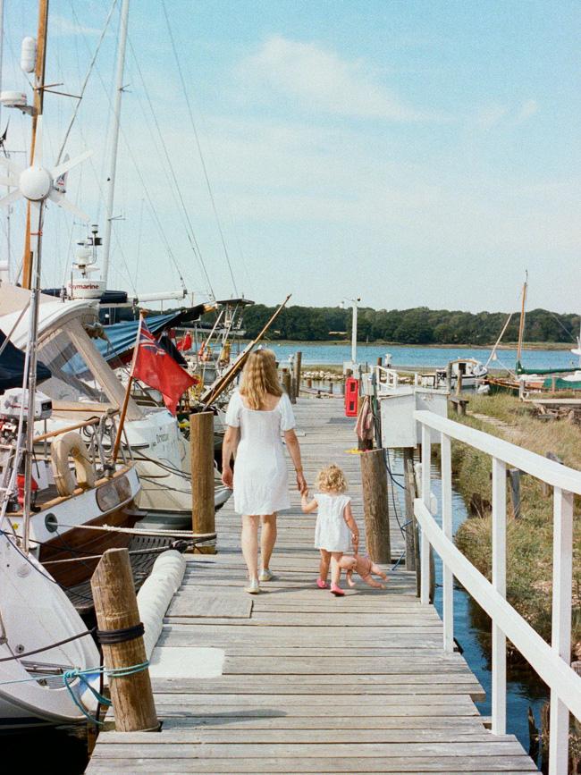 A woman and a young girl walk hand in hand along a wooden dock, surrounded by sailboats and a serene body of water. The atmosphere is peaceful and idyllic, with a sense of.