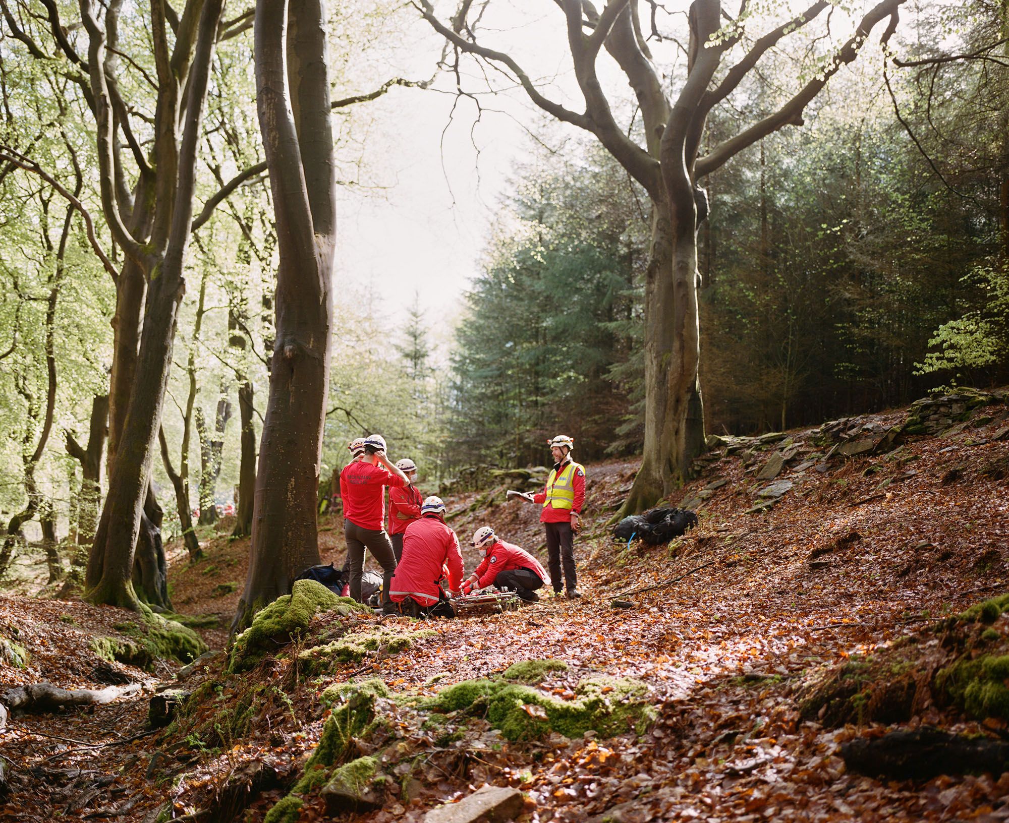 A group of people in red jackets and helmets are gathered around a person on the ground in a forest.