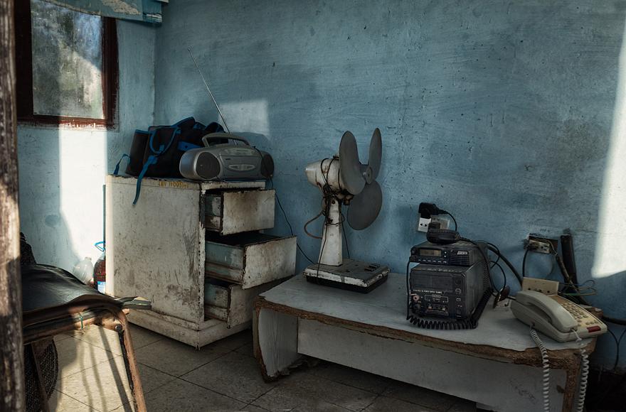 A room with a table, a chair, and a few objects on the table, including a radio, a fan, and a few other items.