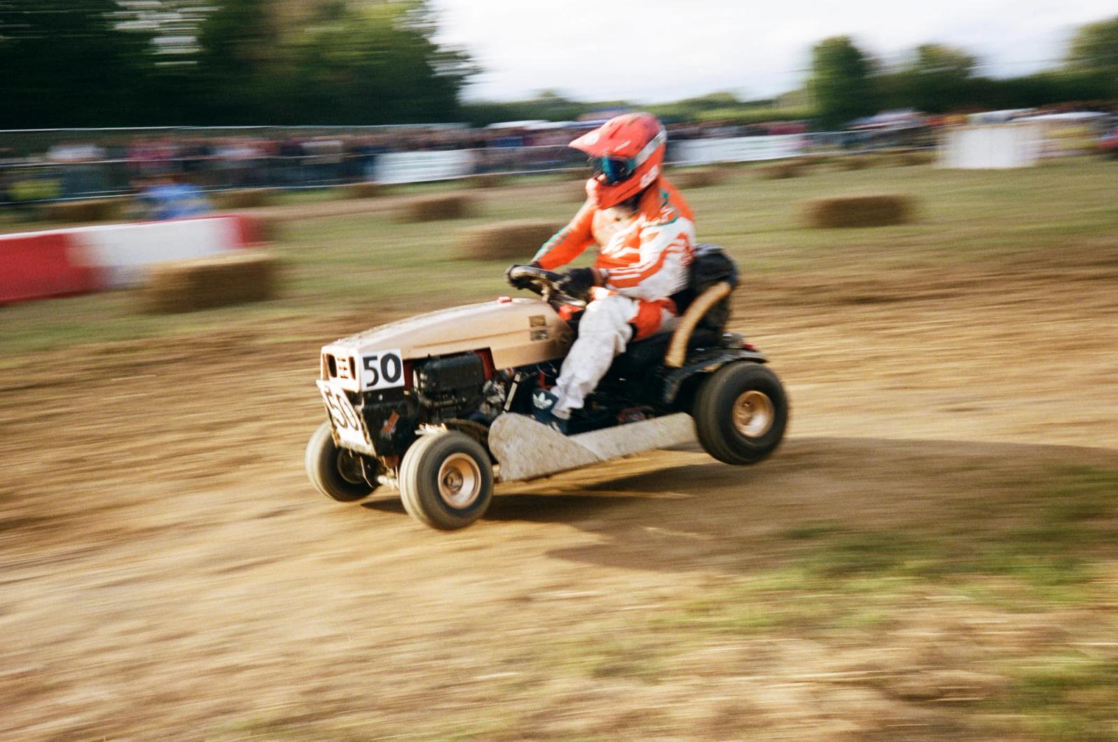 A person in an orange and white racing suit and helmet is driving a small, off-road, lawn mower-like vehicle with a number 50 on the front. The person is wearing a helmet with a.