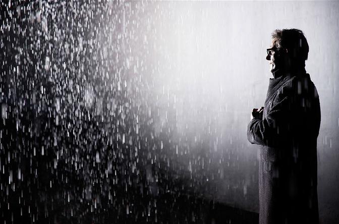 A man in a long, dark coat stands in a room filled with rain, looking up at the ceiling.