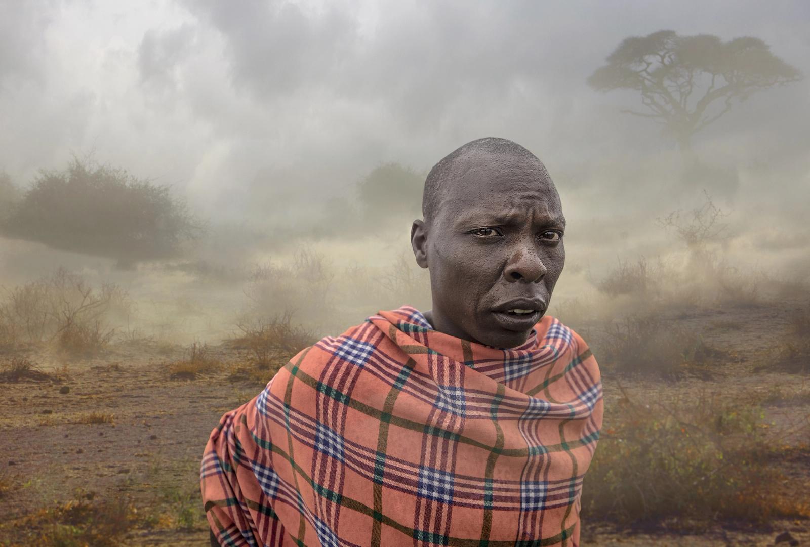 A Maasai tribesman in a traditional red and blue plaid shawl, standing in a dry, grassy savannah landscape.