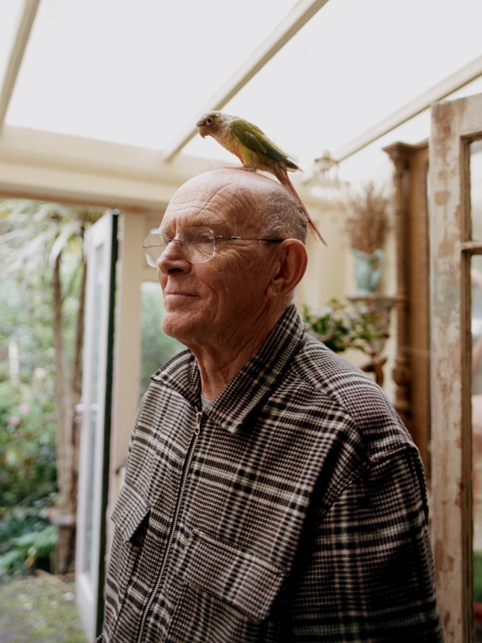 An older man with a green and white parrot perched on his head, wearing a plaid shirt and glasses, in a sunroom with a garden view.