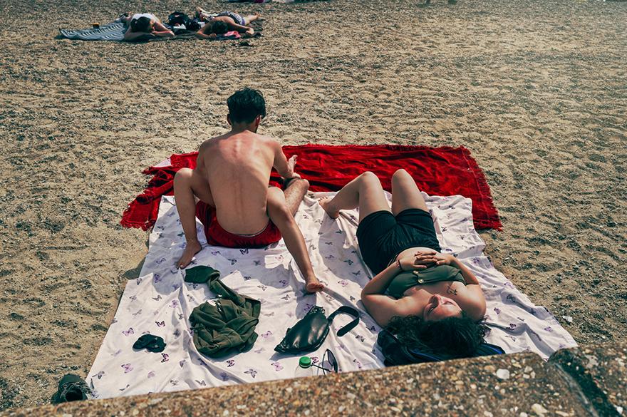 A group of people are sunbathing on a beach, with some wearing swimsuits and others wearing beach towels.