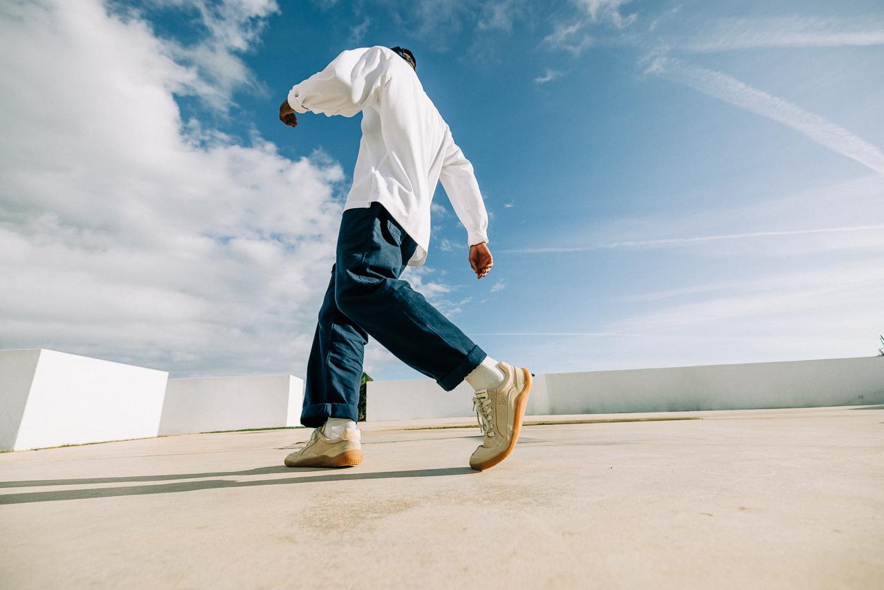 A man in a white shirt and blue jeans walking on a rooftop with a blue sky and clouds in the background.