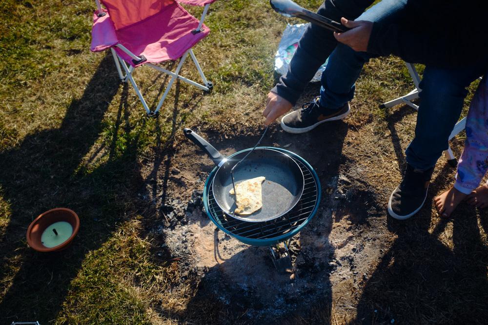 A person is cooking a tortilla on a portable grill in a grassy area. The person is sitting in a pink camping chair, holding a spatula, and appears to be enjoying the warm, sunny.