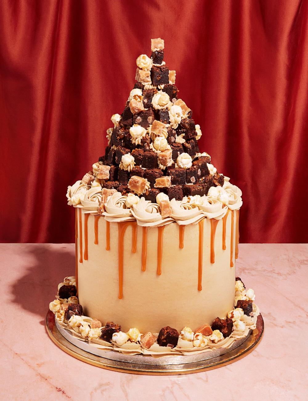 A large, multi-tiered cake with a pyramid-shaped brownie and caramel sauce topping, on a pink marble table in front of a red curtain.