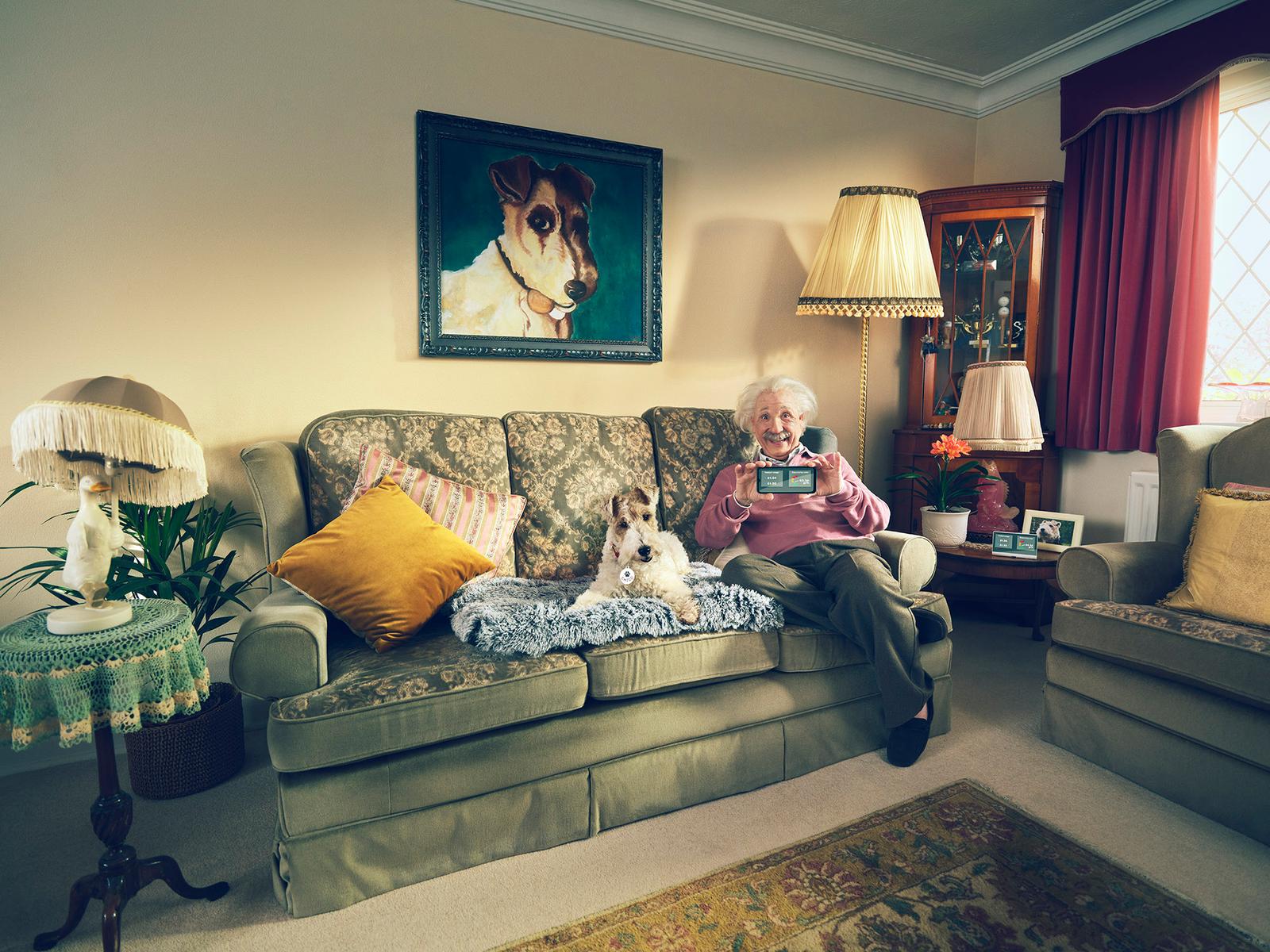 An elderly woman sits on a couch with two small white dogs, surrounded by various decorative items and a painting of a dog on the wall.