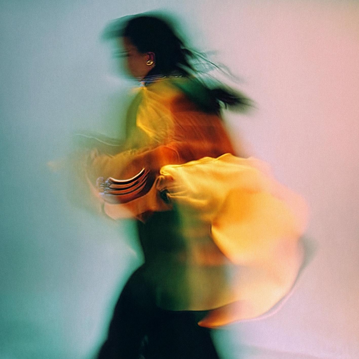 A woman in a flowing, long-sleeved, yellow dress with a high collar and a full, flared skirt, dancing in a blurred, dreamy, ethereal environment.