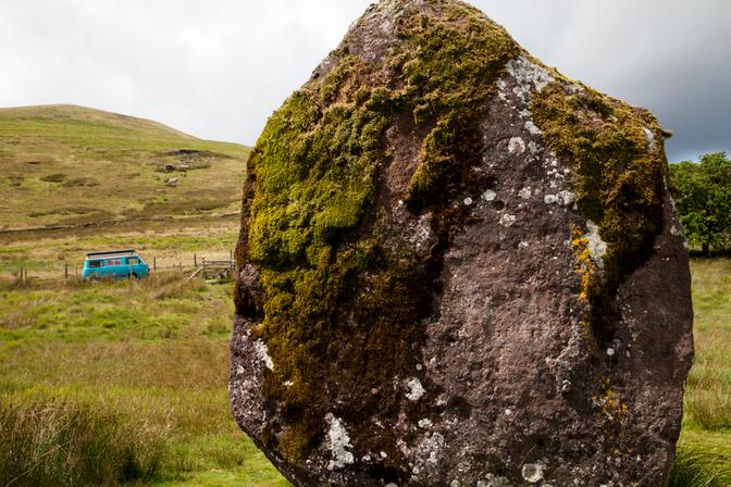 A large, moss-covered rock in a rural landscape, with a small blue vehicle in the background.