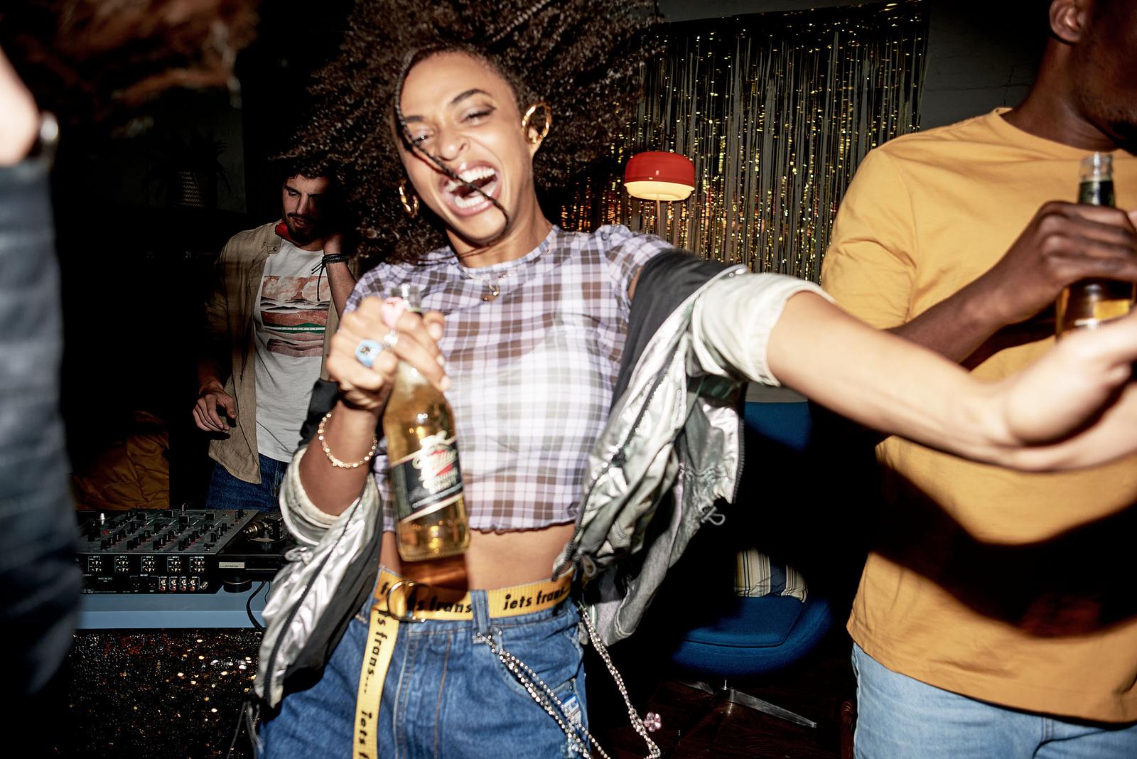 A woman dances while holding a miller beer bottle in a dimly lit room during a house party.