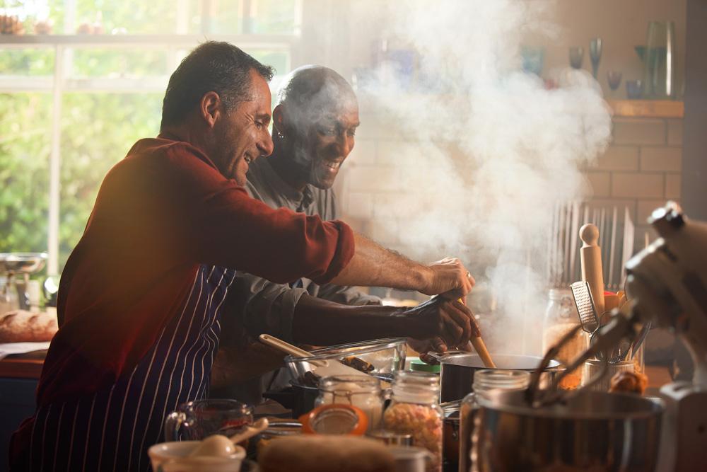 Two men, one wearing a red shirt and the other a grey shirt, cooking in a kitchen with steam rising from a pot.