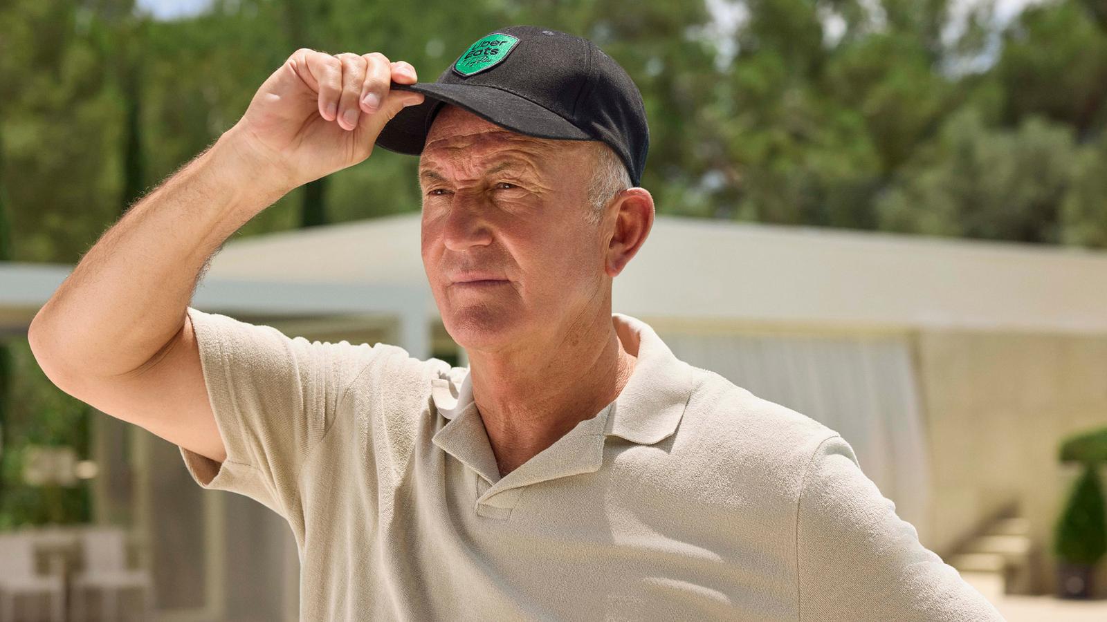 A man wearing a black baseball cap with a green logo on the front, standing in front of a blurred background.